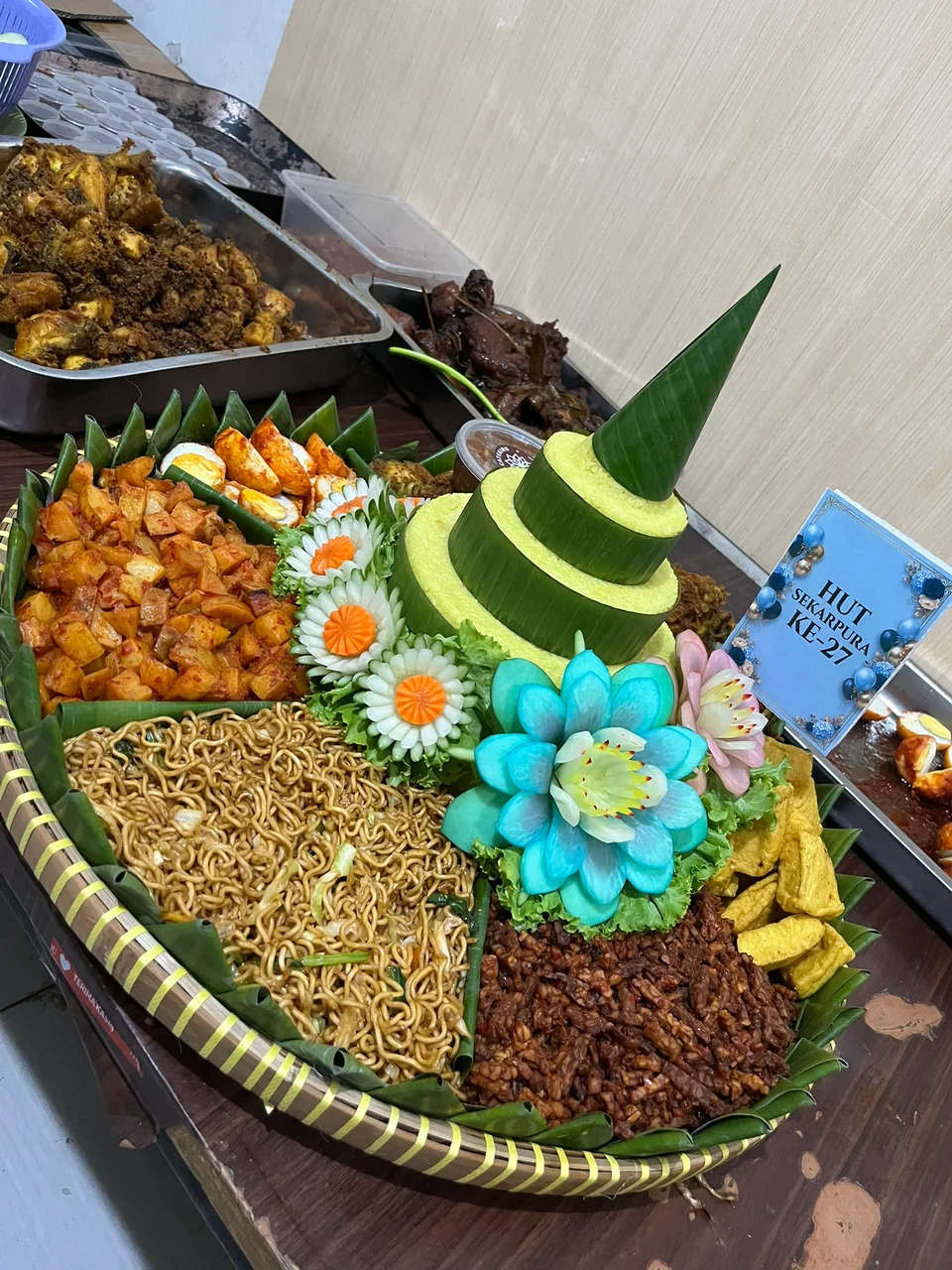 Tumpeng kuning bertingkat dengan hiasan bunga biru dan aneka lauk di tampah, untuk perayaan ulang tahun ke-27.