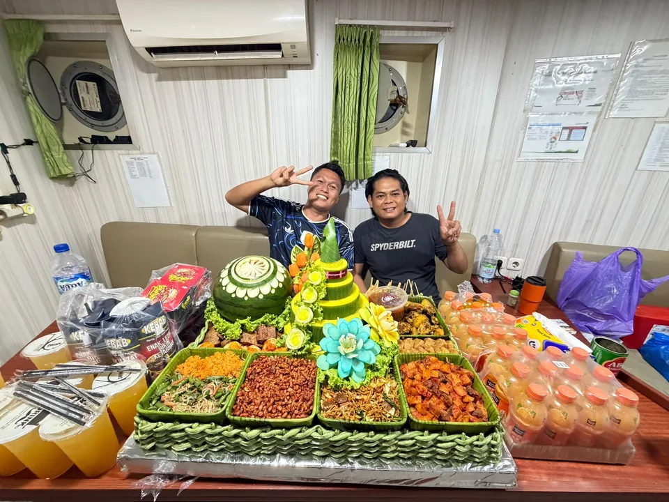 Dua orang berpose di belakang tumpeng bertingkat, ukiran semangka, aneka lauk, dan minuman di atas meja.