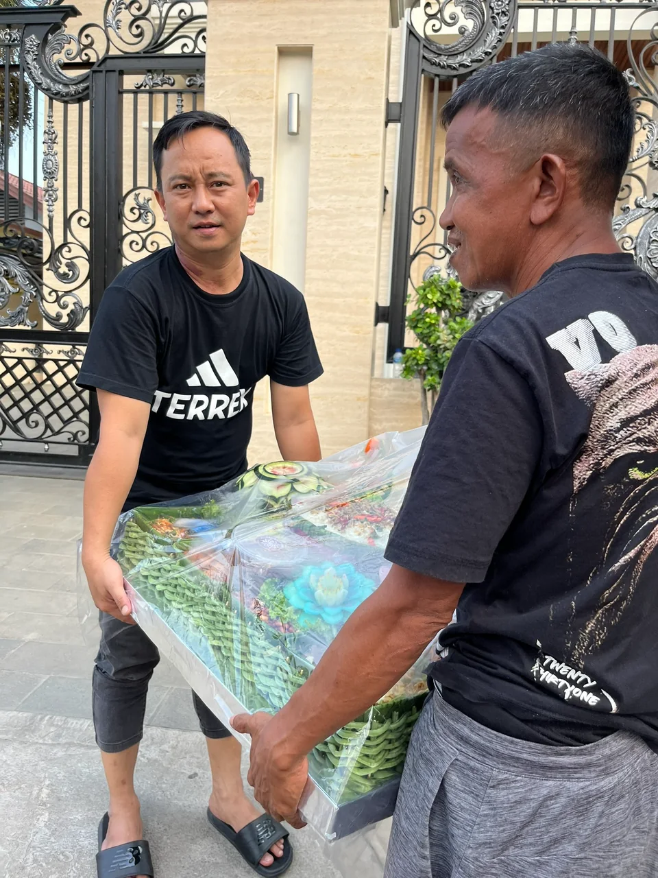 Dua orang membawa bersama tampah tumpeng besar yang masih terbungkus plastik di depan gerbang rumah.