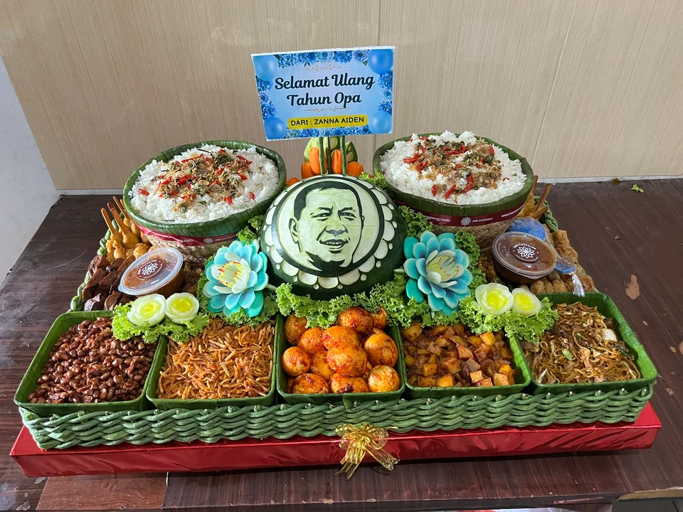 Hantaran ulang tahun Opa dengan dua bakul nasi, ukiran wajah pada semangka, hiasan bunga, dan lauk di tampah besar.